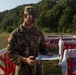 A U.S. Army soldier in OCP (Operational Camouflage Pattern) uniform is reviewing a document or map while standing among red-and-white barrier structures in an outdoor field setting. The soldier wears a name tape, rank insignia, and a U.S. flag patch on the left shoulder, indicating active duty status. The setting appears to be a training exercise or operational planning area with wooded hills in the background.