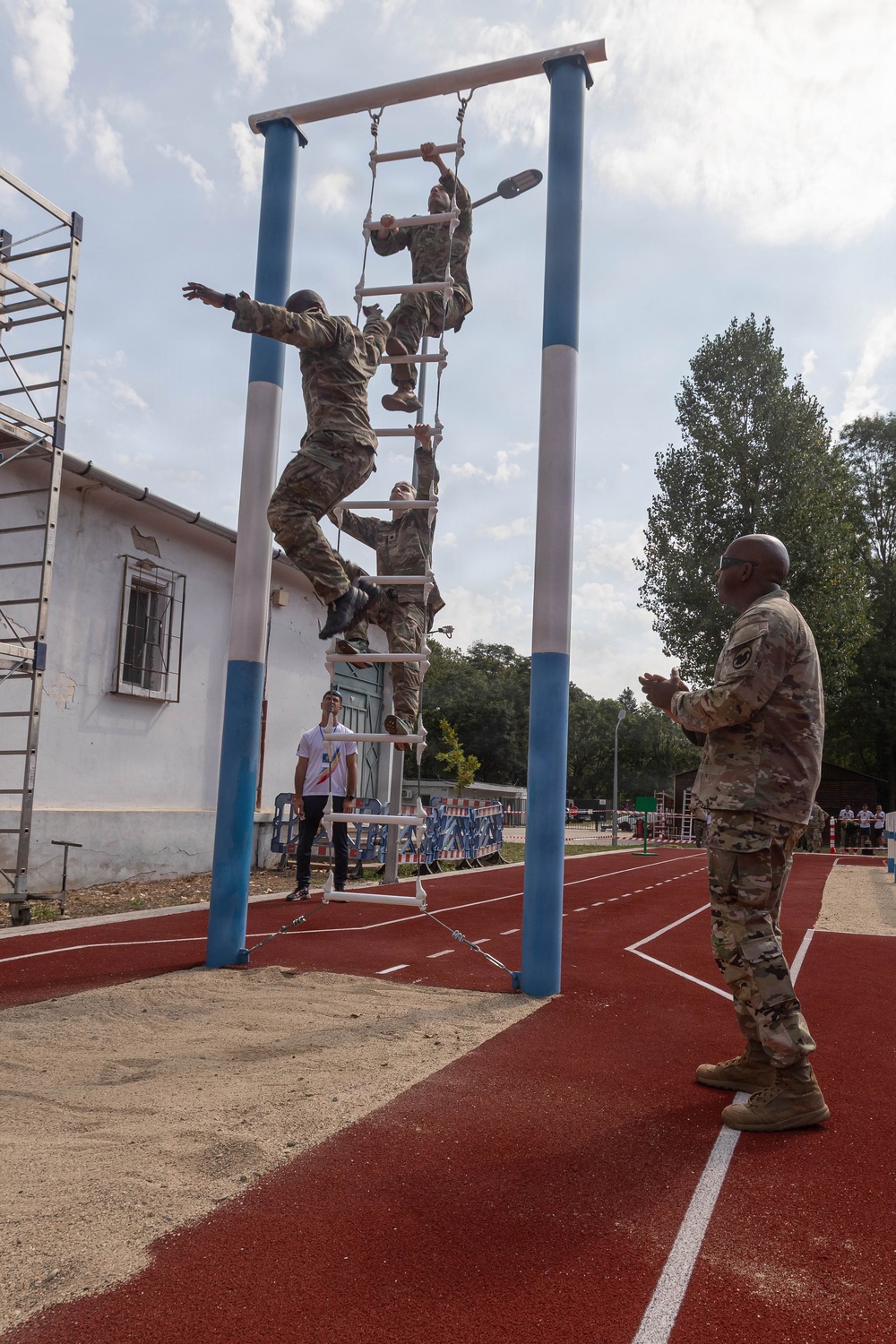 Multiple soldiers in camouflage uniforms are climbing a vertical rope ladder obstacle on what appears to be a military training course. A senior soldier in camouflage stands to the right observing and appears to be clapping or gesturing, while a civilian in a white shirt watches from the background. The setting includes a red rubberized running track, blue and white painted support poles, and a sandy landing pit beneath the obstacle.