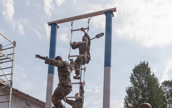 Multiple soldiers in camouflage uniforms are climbing a vertical rope ladder obstacle on what appears to be a military training course. A senior soldier in camouflage stands to the right observing and appears to be clapping or gesturing, while a civilian in a white shirt watches from the background. The setting includes a red rubberized running track, blue and white painted support poles, and a sandy landing pit beneath the obstacle.