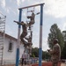 Multiple soldiers in camouflage uniforms are climbing a vertical rope ladder obstacle on what appears to be a military training course. A senior soldier in camouflage stands to the right observing and appears to be clapping or gesturing, while a civilian in a white shirt watches from the background. The setting includes a red rubberized running track, blue and white painted support poles, and a sandy landing pit beneath the obstacle.