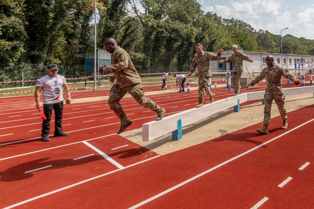 Military personnel in camouflage uniforms are participating in a long jump or broad jump athletic event on a red running track. One soldier is captured mid-air during a jump over a sand pit with blue markers, while others observe or prepare nearby. A civilian official in a white shirt stands to the left, likely serving as a judge or timekeeper.