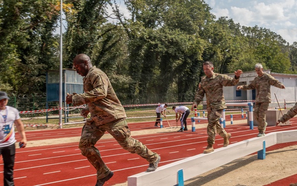 Military personnel in camouflage uniforms are participating in a long jump or broad jump athletic event on a red running track. One soldier is captured mid-air during a jump over a sand pit with blue markers, while others observe or prepare nearby. A civilian official in a white shirt stands to the left, likely serving as a judge or timekeeper.