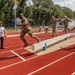 Military personnel in camouflage uniforms are participating in a long jump or broad jump athletic event on a red running track. One soldier is captured mid-air during a jump over a sand pit with blue markers, while others observe or prepare nearby. A civilian official in a white shirt stands to the left, likely serving as a judge or timekeeper.