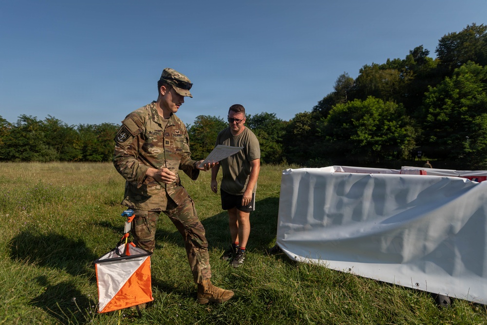 A U.S. Army soldier in full OCP (Operational Camouflage Pattern) uniform reviews a document with a civilian or off-duty military individual in a grassy field. An orange-and-white orienteering marker is visible on the ground nearby, along with a large white panel structure, suggesting a land navigation or orienteering exercise. The setting appears to be a training area with tree lines in the background under clear blue skies.