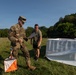 A U.S. Army soldier in full OCP (Operational Camouflage Pattern) uniform reviews a document with a civilian or off-duty military individual in a grassy field. An orange-and-white orienteering marker is visible on the ground nearby, along with a large white panel structure, suggesting a land navigation or orienteering exercise. The setting appears to be a training area with tree lines in the background under clear blue skies.