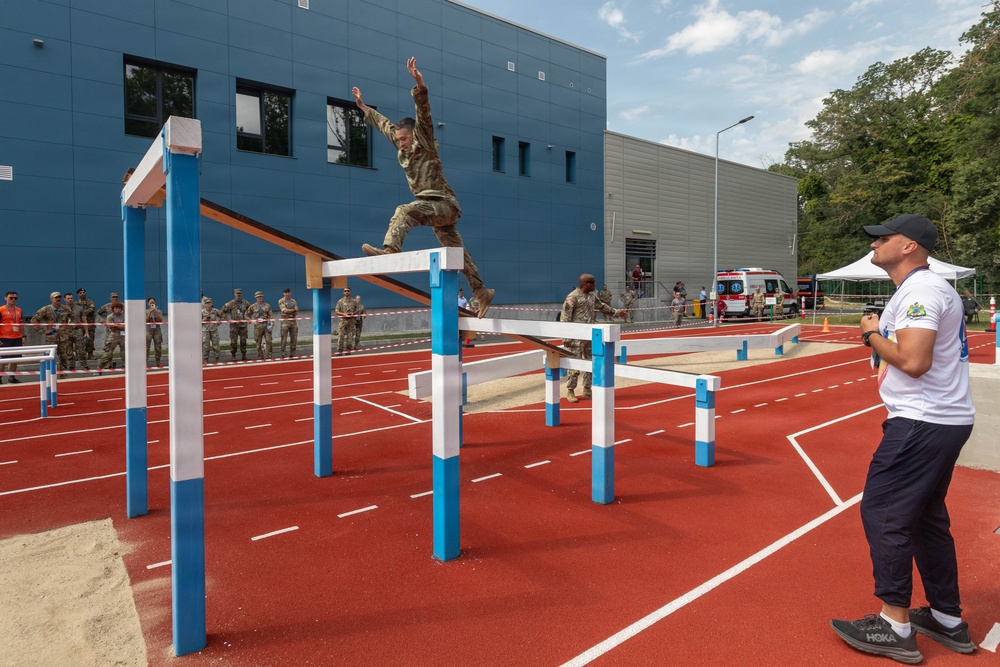 A soldier in camouflage uniform is navigating an outdoor military obstacle course, leaping over a horizontal bar structure on a red rubberized track surface. A civilian official in a white shirt and dark cap observes and appears to be timing or judging the performance. A group of uniformed military personnel watches from behind a rope barrier in the background.