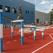 A soldier in camouflage uniform is navigating an outdoor military obstacle course, leaping over a horizontal bar structure on a red rubberized track surface. A civilian official in a white shirt and dark cap observes and appears to be timing or judging the performance. A group of uniformed military personnel watches from behind a rope barrier in the background.