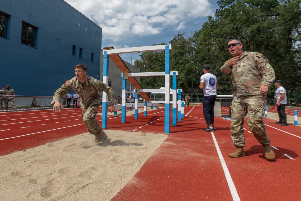 Two soldiers in camouflage uniforms are participating in a military athletic competition on a red running track. One soldier appears to have just completed a long jump, landing in a sand pit, while a second soldier nearby appears to be coaching or observing. The event takes place at an outdoor sports facility adjacent to a modern blue building, with other personnel visible in the background.