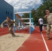 Two soldiers in camouflage uniforms are participating in a military athletic competition on a red running track. One soldier appears to have just completed a long jump, landing in a sand pit, while a second soldier nearby appears to be coaching or observing. The event takes place at an outdoor sports facility adjacent to a modern blue building, with other personnel visible in the background.