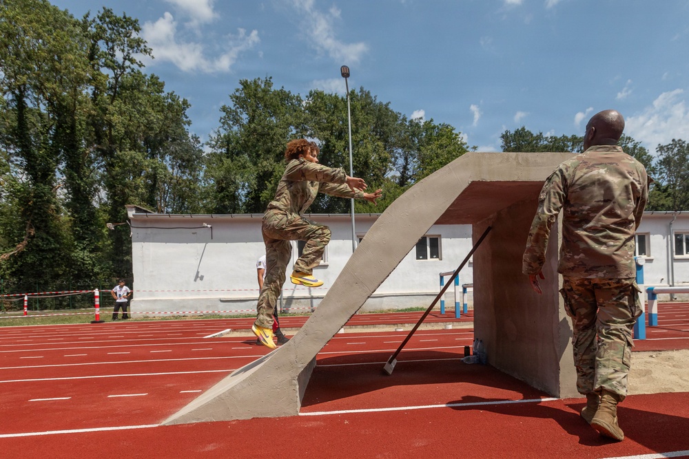 Two military personnel in camouflage uniforms are on a red athletic track, with one soldier actively running up or jumping over a concrete obstacle structure. A second soldier observes from the right side, appearing to supervise or evaluate the activity. The setting appears to be a military physical training or obstacle course exercise conducted at an outdoor sports facility.