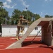 Two military personnel in camouflage uniforms are on a red athletic track, with one soldier actively running up or jumping over a concrete obstacle structure. A second soldier observes from the right side, appearing to supervise or evaluate the activity. The setting appears to be a military physical training or obstacle course exercise conducted at an outdoor sports facility.