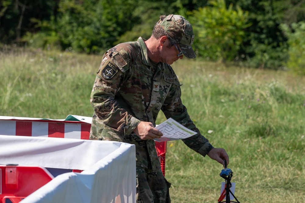 A military personnel wearing OCP (Operational Camouflage Pattern) uniform is examining a document or form near a red and white barrier in an outdoor field setting. The soldier appears to be conducting an administrative or evaluation task, holding papers while handling equipment near what appears to be a checkpoint or staging area. The setting suggests a training exercise or field operation.