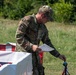 A military personnel wearing OCP (Operational Camouflage Pattern) uniform is examining a document or form near a red and white barrier in an outdoor field setting. The soldier appears to be conducting an administrative or evaluation task, holding papers while handling equipment near what appears to be a checkpoint or staging area. The setting suggests a training exercise or field operation.