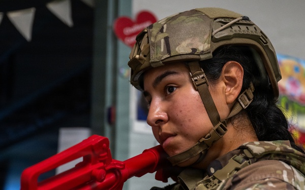 A female military security forces (SF) member in full tactical gear, including a camouflage uniform and combat helmet, holds a bright red training/dummy weapon. The setting appears to be an indoor facility, possibly a school or community center, with decorative banners and a heart-shaped decoration visible in the background. The red weapon is clearly a non-functional training replica, used for drills or demonstrations.