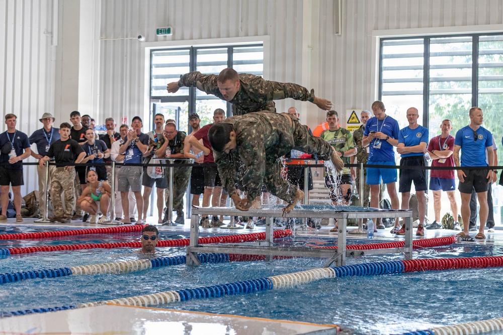 Military personnel in camouflage uniforms are performing a pool obstacle course event at an indoor aquatic facility, with one soldier diving into the water over a metal barrier while another prepares to follow. A large crowd of spectators, many wearing lanyards and badges suggesting an official competition, lines the poolside barrier. The event appears to be a military swimming competition or fitness challenge.