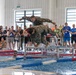 Military personnel in camouflage uniforms are performing a pool obstacle course event at an indoor aquatic facility, with one soldier diving into the water over a metal barrier while another prepares to follow. A large crowd of spectators, many wearing lanyards and badges suggesting an official competition, lines the poolside barrier. The event appears to be a military swimming competition or fitness challenge.
