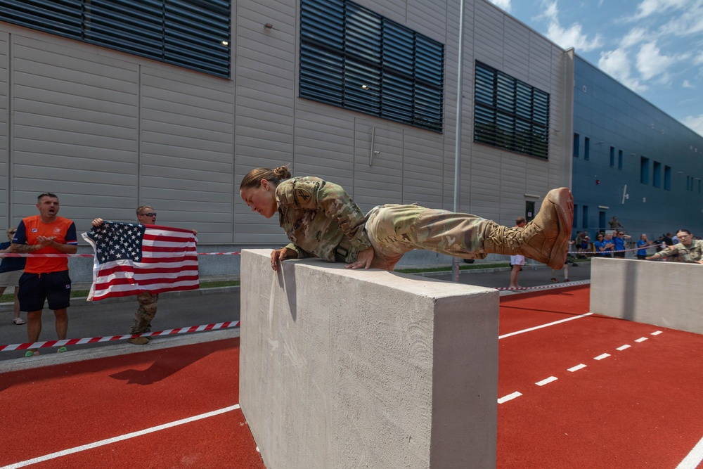A soldier in full camouflage uniform is performing an obstacle course challenge, scaling or vaulting over a large concrete block on a red athletic track. Spectators, including one holding an American flag, cheer from behind a barrier tape. The setting appears to be a military fitness competition or international military sports event.