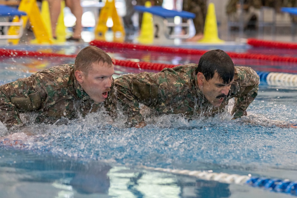 Two individuals wearing matching camouflage military uniforms are swimming in an indoor pool, appearing to perform a military water training exercise. Both are fully clothed in combat fatigues while swimming, suggesting this is a formal military fitness or qualification assessment. The setting is an organized indoor aquatic facility with lane markers and safety cones visible in the background.