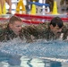 Two individuals wearing matching camouflage military uniforms are swimming in an indoor pool, appearing to perform a military water training exercise. Both are fully clothed in combat fatigues while swimming, suggesting this is a formal military fitness or qualification assessment. The setting is an organized indoor aquatic facility with lane markers and safety cones visible in the background.