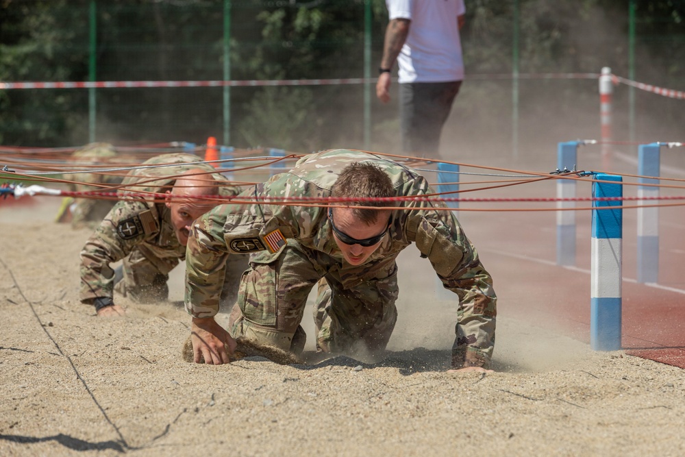 Two soldiers in camouflage uniforms are crawling under a low wire obstacle course through a sandy pit, generating dust as they move. Both wear multicam pattern uniforms with visible unit patches including US flag patches. A supervisor or observer in civilian clothing stands in the background overseeing the exercise.