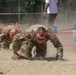 Two soldiers in camouflage uniforms are crawling under a low wire obstacle course through a sandy pit, generating dust as they move. Both wear multicam pattern uniforms with visible unit patches including US flag patches. A supervisor or observer in civilian clothing stands in the background overseeing the exercise.
