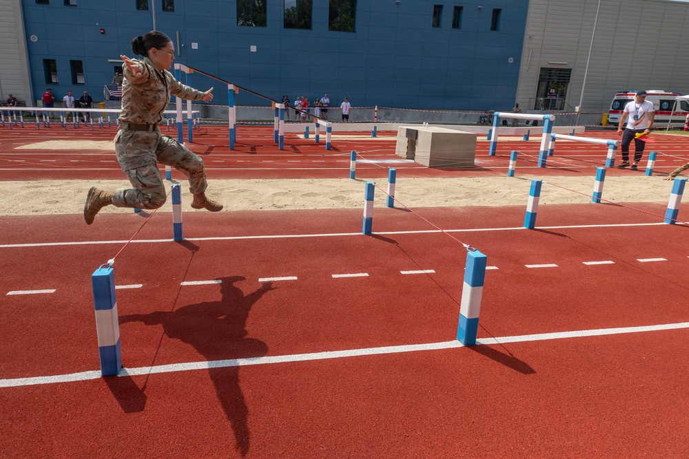 A soldier in camouflage uniform is captured mid-air while jumping over a rope obstacle on a red athletic track during what appears to be a military fitness or obstacle course competition. The course features blue and white striped poles connected by ropes arranged in a grid pattern across the track and a sand pit area. Spectators and officials are visible in the background near a modern blue building.