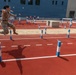 A soldier in camouflage uniform is captured mid-air while jumping over a rope obstacle on a red athletic track during what appears to be a military fitness or obstacle course competition. The course features blue and white striped poles connected by ropes arranged in a grid pattern across the track and a sand pit area. Spectators and officials are visible in the background near a modern blue building.