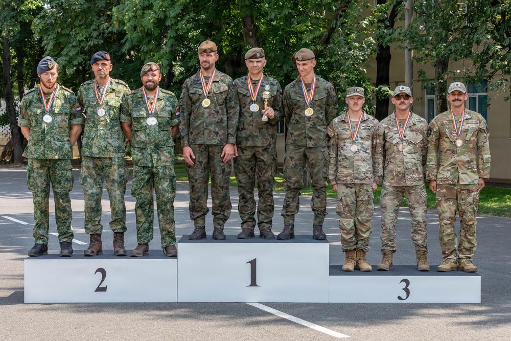 A military awards ceremony podium photograph showing nine soldiers standing on a three-tiered competition podium numbered 1, 2, and 3. The soldiers are wearing different national military uniforms and are displaying medals around their necks, with the first-place team also holding a small trophy. The setting appears to be an outdoor military installation with trees and a building visible in the background.
