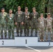 A military awards ceremony podium photograph showing nine soldiers standing on a three-tiered competition podium numbered 1, 2, and 3. The soldiers are wearing different national military uniforms and are displaying medals around their necks, with the first-place team also holding a small trophy. The setting appears to be an outdoor military installation with trees and a building visible in the background.
