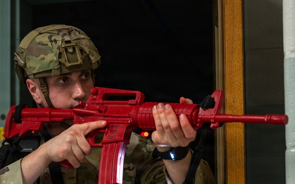 A military or law enforcement trainee in full combat gear is conducting a building clearing exercise using a bright red training weapon (inert replica). The red-colored firearm is a standard training aid used to distinguish simulated weapons from live ones during drills. The setting appears to be an indoor facility, likely a school or office building, used for active shooter or tactical response training.