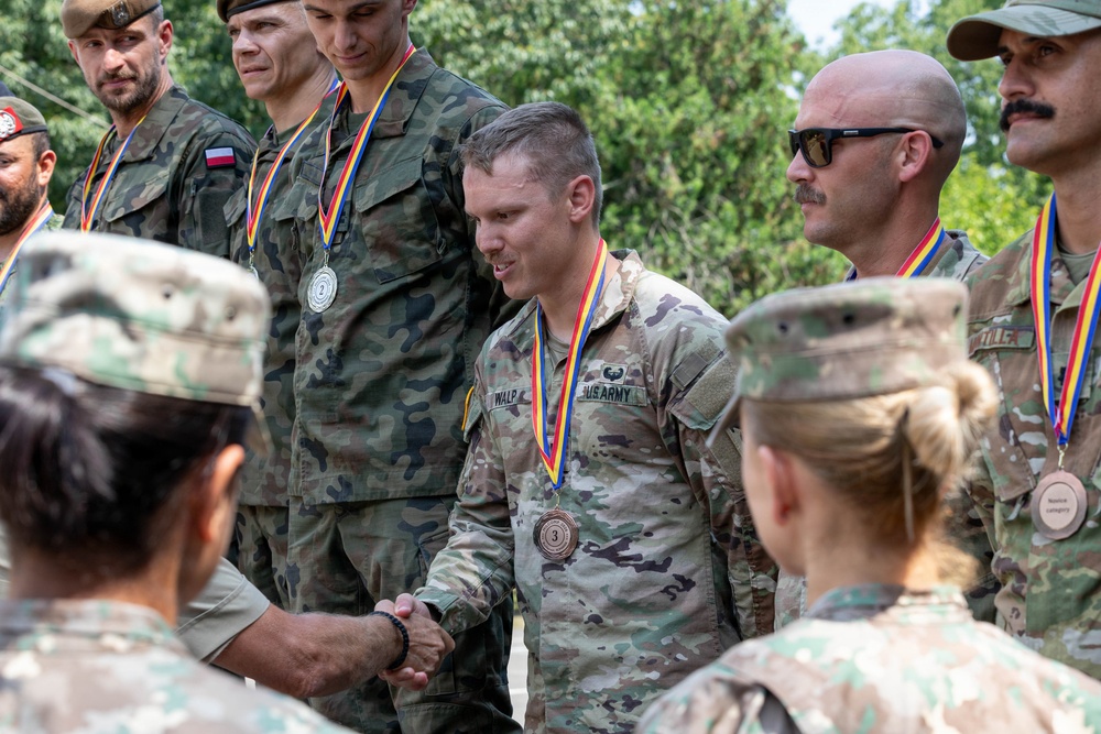 A military awards ceremony featuring soldiers from multiple nations, including US Army personnel and Polish military members, receiving medals with red, yellow, and blue ribbons. The central figure, identified by name tape as 'WALP' from the US Army, is wearing a bronze (3rd place) medal and shaking hands with another soldier. The setting appears to be an outdoor multinational military competition or exercise.