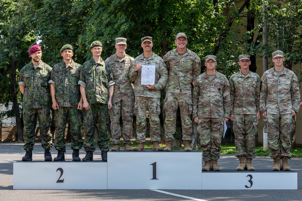 A military awards ceremony showing soldiers standing on a competition podium with positions 1, 2, and 3 marked. The first-place team, wearing US Army OCP (Operational Camouflage Pattern) uniforms, is holding a diploma or certificate. The second-place team appears to be wearing Eastern European military uniforms, likely Czech or Slovak, while the third-place team also wears US Army uniforms.