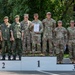 A military awards ceremony showing soldiers standing on a competition podium with positions 1, 2, and 3 marked. The first-place team, wearing US Army OCP (Operational Camouflage Pattern) uniforms, is holding a diploma or certificate. The second-place team appears to be wearing Eastern European military uniforms, likely Czech or Slovak, while the third-place team also wears US Army uniforms.