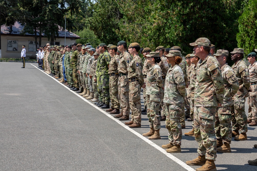 A multinational military formation is standing at attention during what appears to be a formal parade or ceremony on a paved surface. Personnel are wearing various camouflage uniforms representing different nations, with at least two officers in white dress uniforms visible on the far left. The setting appears to be a military installation with trees and a building visible in the background.