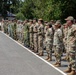 A multinational military formation is standing at attention during what appears to be a formal parade or ceremony on a paved surface. Personnel are wearing various camouflage uniforms representing different nations, with at least two officers in white dress uniforms visible on the far left. The setting appears to be a military installation with trees and a building visible in the background.