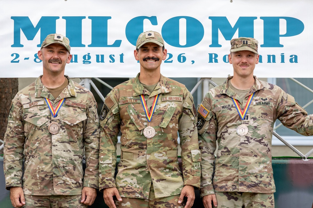 Three U.S. military personnel are posing in front of a 'MILCOMP' banner dated August 2-8, 2026, held in Romania. All three are wearing camouflage uniforms and displaying bronze medals with red, yellow, and blue ribbon lanyards, indicating a third-place finish. The individuals represent different branches: U.S. Army (left and right) and U.S. Air Force (center).