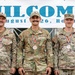 Three U.S. military personnel are posing in front of a 'MILCOMP' banner dated August 2-8, 2026, held in Romania. All three are wearing camouflage uniforms and displaying bronze medals with red, yellow, and blue ribbon lanyards, indicating a third-place finish. The individuals represent different branches: U.S. Army (left and right) and U.S. Air Force (center).