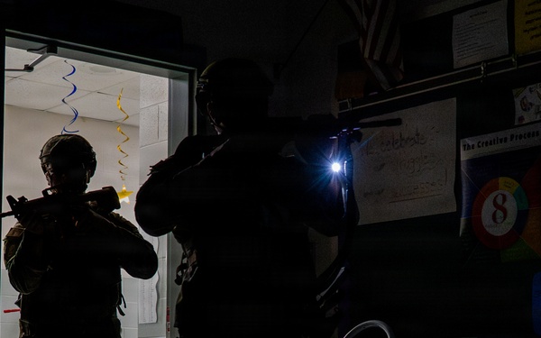 Two heavily equipped tactical personnel in full gear and helmets are conducting a room-clearing operation inside what appears to be a school classroom. One operator is positioned in the doorway while the second uses a weapon-mounted flashlight to illuminate the darkened room. The setting shows clear indicators of a school environment, suggesting this is a law enforcement or military active-shooter response drill or training exercise.