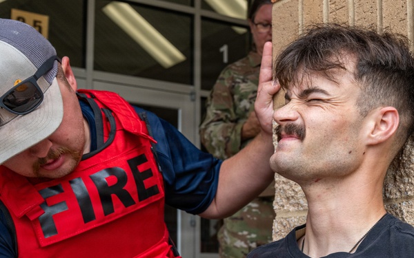 A firefighter/first responder wearing a red 'FIRE' vest and gray cap is providing medical assistance to a seated male casualty who appears to be in distress. The responder is examining the casualty's head/face area while applying pressure to what appears to be a wound on the chest. Military personnel in camouflage uniforms are visible in the background, suggesting this may be a mass casualty training exercise.