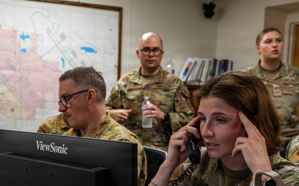 Military personnel in camouflage uniforms are working in what appears to be a command or operations center. A female soldier in the foreground, identified by a name tape reading 'STABELL', is on a corded telephone while pressing her finger to her temple in concentration. Several other uniformed personnel are visible in the background near computers and a wall-mounted map.