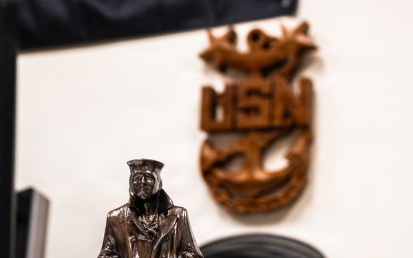 A bronze-finish miniature replica statue of 'The Lone Sailor' from the United States Navy Memorial is displayed on a desk. The figurine depicts a sailor in uniform standing in a relaxed pose on a wooden base with a black and gold engraved plaque. In the background, a wooden USN (United States Navy) Chief Petty Officer anchor insignia is mounted on the wall.