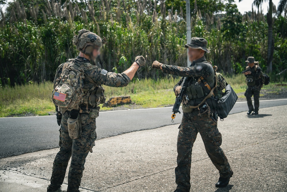 Two armed military personnel in camouflage uniforms are performing a fist bump on a paved road in a tropical environment. The soldier on the left displays a U.S. flag patch on their right shoulder, while the soldier on the right carries a rifle and tactical gear. A third uniformed individual is visible in the background.