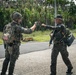 Two armed military personnel in camouflage uniforms are performing a fist bump on a paved road in a tropical environment. The soldier on the left displays a U.S. flag patch on their right shoulder, while the soldier on the right carries a rifle and tactical gear. A third uniformed individual is visible in the background.