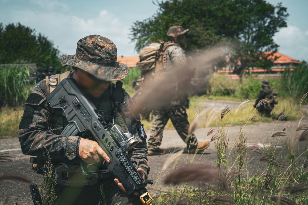 The image depicts multiple military personnel in camouflage uniforms conducting what appears to be a tactical field exercise or patrol operation in a semi-urban, vegetated environment. The foreground soldier is armed with a bullpup-style assault rifle and wearing a boonie hat with digital camouflage pattern. A smoke or dust cloud is visible near the center of the frame, suggesting active movement or a smoke grenade deployment.