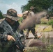 The image depicts multiple military personnel in camouflage uniforms conducting what appears to be a tactical field exercise or patrol operation in a semi-urban, vegetated environment. The foreground soldier is armed with a bullpup-style assault rifle and wearing a boonie hat with digital camouflage pattern. A smoke or dust cloud is visible near the center of the frame, suggesting active movement or a smoke grenade deployment.