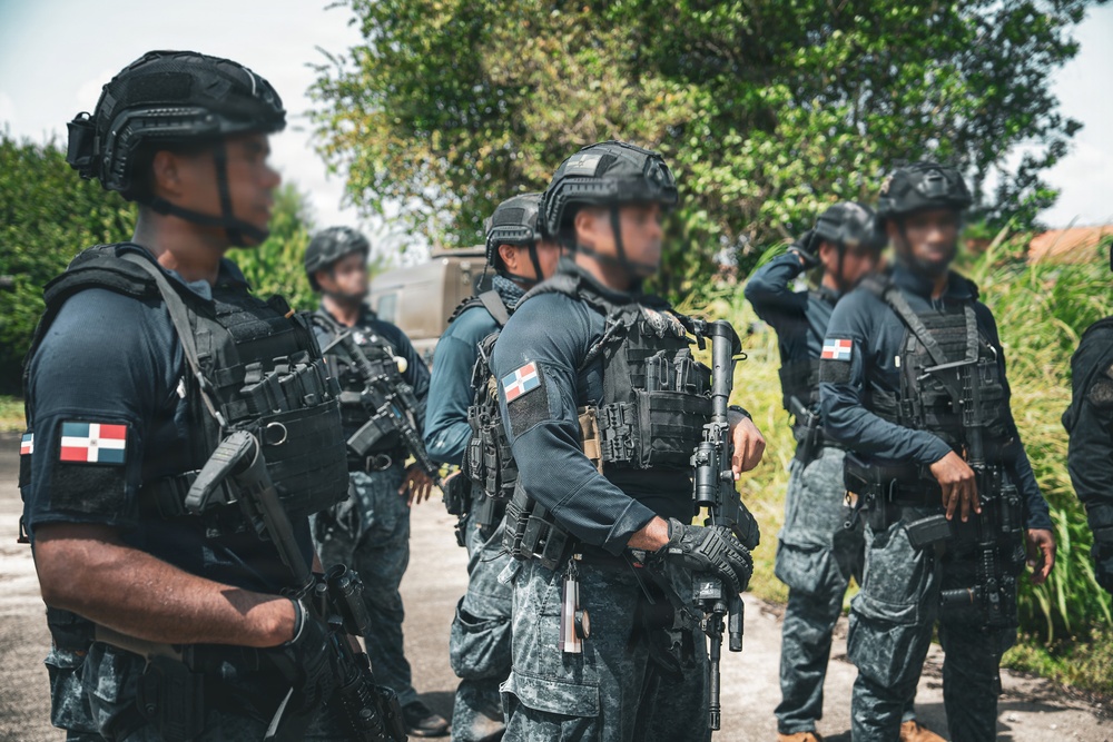 A group of heavily armed tactical/special operations personnel are assembled outdoors in a formation or briefing posture. Multiple individuals are wearing Dominican Republic flag patches on their uniforms, identifying them as Dominican security forces. They are equipped with full tactical gear including body armor, helmets, and assault rifles.