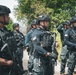 A group of heavily armed tactical/special operations personnel are assembled outdoors in a formation or briefing posture. Multiple individuals are wearing Dominican Republic flag patches on their uniforms, identifying them as Dominican security forces. They are equipped with full tactical gear including body armor, helmets, and assault rifles.