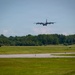 A four-engine turboprop military transport aircraft, consistent with a C-130 Hercules, is captured on final approach or low-level pass over a runway. The aircraft is viewed head-on at low altitude above a paved airstrip surrounded by green grass and a treeline in the background. The image was taken from ground level at a military or civilian airfield during daytime under partly cloudy skies.