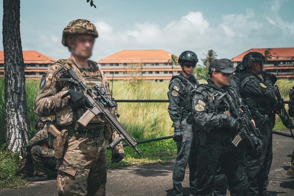 The image shows a group of armed military or law enforcement personnel in an outdoor setting near a building with red-tiled roofs. One individual in the foreground wears multicam camouflage and carries a rifle with optics, while others in the background wear all-black tactical uniforms and helmets. The scene appears to depict a joint training exercise or operational briefing between different units or nationalities.