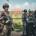 The image shows a group of armed military or law enforcement personnel in an outdoor setting near a building with red-tiled roofs. One individual in the foreground wears multicam camouflage and carries a rifle with optics, while others in the background wear all-black tactical uniforms and helmets. The scene appears to depict a joint training exercise or operational briefing between different units or nationalities.